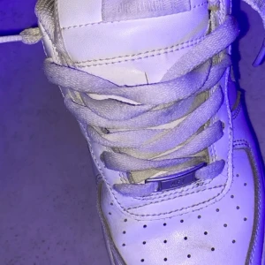 Vita Nike Air Force sneakers - Klassiska vita Nike Air Force sneakers med snörning och perforerade detaljer på tån. Skorna har en robust sula och Nike-logga på hälen. Perfekta för en stilren och sportig look.