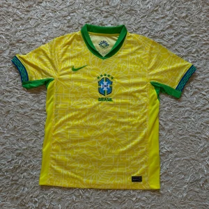 BRASIL FOTBOLLSJERSEY från Nike - Säljer en gul Brasil fotbollströja från Nike med gröna detaljer och ett subtilt mönster över hela tröjan. Tröjan har korta ärmar och det ikoniska CBF-emblemet på bröstet. Perfekt för alla fotbollsfans som vill visa sitt stöd för Brasilien! 🇧🇷