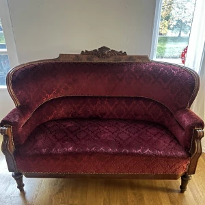 Röd viktoriansk soffa - !!Man kan använda sig utav tipptapp-appen för att leverera hem sig soffan!!  Den här soffan är ett utsökt exempel på antik möbelkonst, troligtvis från slutet av 1800-talet eller början av 1900-talet, ifrån Stockholm, med en stark influens av viktoriansk eller barock stil. Den robusta träramen är vackert utskuren med dekorativa detaljer, särskilt framträdande på den övre ryggen, vilket indikerar skickligt hantverk. Träet har en djup, mörk finish som kontrasterar stilfullt mot den rika klädseln.