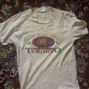 Vintage tshirt - Säljer en beige t-shirt med ett coolt tryck av en sol och texten 'Toronto'. T-shirten har korta ärmar och en rund halsringning. Perfekt för dig som gillar unika tryck och vill ha något annorlunda i garderoben.
