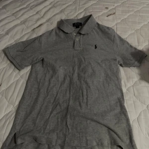 Grå pikétröja från Ralph Lauren - Säljer en klassisk grå pikétröja från Ralph Lauren med korta ärmar och broderad logga på bröstet. Perfekt för en stilren look. Tröjan har en krage och knappar framtill.