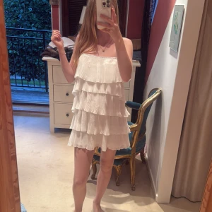 Vit volangklänning med plissering - Supersöt vit kortklänning med flera lager volanger och plisserat tyg. Axelbandslös modell som ger en luftig och lekfull känsla, perfekt för varma dagar. Klänningen har en loose passform och är tillverkad i ett lätt material. Aldrig använd❤️