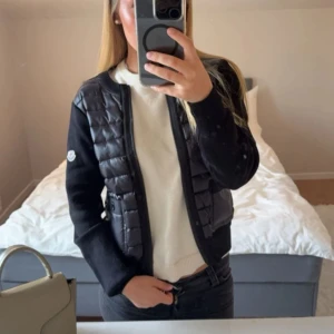 Moncler cardigan - Snygg svart dunväst från Moncler med quiltad framsida och två fickor med knappar. Västen har stickade ärmar och rygg, rund halsringning och dragkedja framtill. Perfekt för lager-på-lager och har en clean, sportig look.