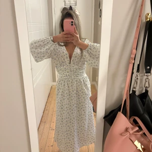 Långklänning med v-ringning från Gina tricot storlek xs - Superfin vit långklänning med små blå blommor, djup v-ringning och markerad midja. Klänningen är köpt på Gina tricot och är som en adoore klänning. Den är endast använd en gång förra året! Perfekt för sommarens alla tillfällen och ger en fräsch, feminin look.