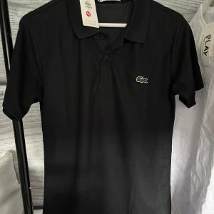 Lacoste piké - Klassisk svart pikétröja från Lacoste i regular fit med korta ärmar och ikoniska krokodilloggan på bröstet. Tröjan har krage och knäppning framtill. Perfekt för en clean och stilren look. Sprillans ny🏝️