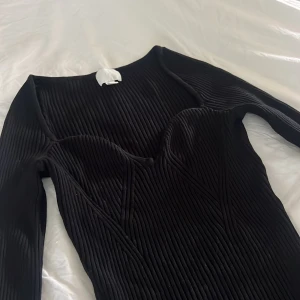 Svart ribbad klänning från H&M - Svart, tight ribbad topp från H&M med lång ärm och markerad byst. Toppen har en snygg, figurnära passform och är tillverkad i stretchigt material som sitter skönt på kroppen. Perfekt för dig som gillar minimalistisk och stilren look.