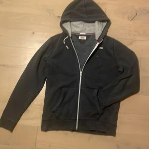 Mörkblå hoodie från Tommy Jeans - Säljer en snygg mörkblå hoodie från Tommy Jeans med dragkedja och justerbar huva. Den har en liten logotyp på bröstet. Perfekt för en avslappnad stil. Storlek står L passar även M