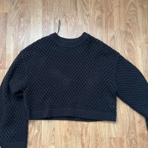 Svart croppad stickad tröja H&M - Säljer en svart croppad stickad tröja från H&M med hålmönster. Tröjan har lång ärm, ribbade muddar och rund halsringning. Perfekt för lager-på-lager och ger en cool vibe till din outfit.