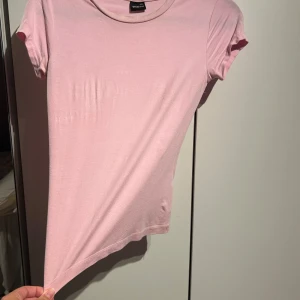 Ljusrosa t-shirt från Gina Tricot - En basic ljusrosa t-shirt från Gina Tricot i mjuk bomull. Modellen är figurnära med korta ärmar och rund halsringning. Perfekt att matcha med jeans eller kjol för en clean och enkel look.