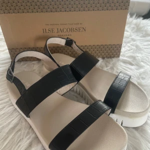 Svarta sandaler från Ilse Jacobsen - Snygga sandaler från Ilse Jacobsen med svarta, breda remmar i skinn med krokopräglad struktur och justerbart spänne. Sulan är beige och har en räfflad design som ger extra höjd och komfort. Perfekta för en trendig sommarlook. I storlek 37 och är i bra skick!👡