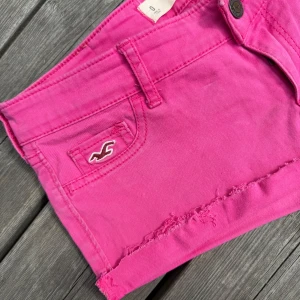 Rosa shorts från Hollister, w 24 - Snygga rosa shorts från Hollister med rå kant nertill och klassisk femficksdesign. Shortsen har låg midja, bälteshällor och en liten broderad logga framtill. Perfekta för sommardagar och ger en cool, avslappnad vibe.