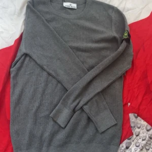 Grå stickad tröja från Stone Island - Säljer en grå stickad tröja från Stone Island med klassisk rund hals och ribbade muddar. Tröjan har den ikoniska Stone Island-loggan på vänster ärm. Perfekt för dig som gillar stilren och enkel design med streetwear-vibe.