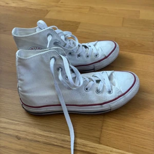 Vita Converse Chuck Taylor All Star - Klassiska vita Converse Chuck Taylor All Star high-tops med röd och blå rand runt sulan. Skorna har snörning, rund tå och är tillverkade i textil med gummisula. Perfekta för en avslappnad och cool stil.