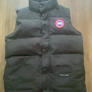 Svart dunväst från Canada Goose - Svart dunväst från Canada Goose med klassisk logga på bröstet. Västen är quiltad och har hög krage samt tryckknappar framtill. Perfekt för lager-på-lager och håller dig varm under kyliga dagar. Snygg och stilren design med diskret ficka framtill. Västen är nästan helt ny, bara använd i ungefär 6-7 dagar.