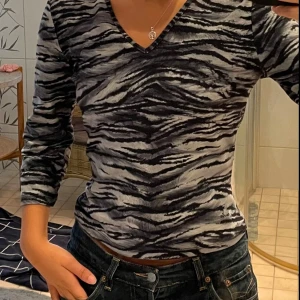 Mönstrad v-ringad tröja - Säljer en snygg zebramönstrad tröja i svart och grått med v-ringning och trekvartsärm. Tröjan har en figurnära passform och är tillverkad i ett mjukt, stretchigt material som känns skönt mot huden. Perfekt för dig som vill sticka ut lite extra.