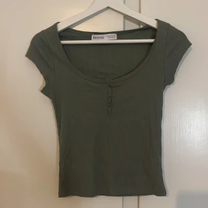 Ribbad grön topp från Bershka XS - Snygg ribbad topp från Bershka i en dämpad grön nyans. Modellen har korta ärmar, rundad halsringning och tre knappar framtill. 