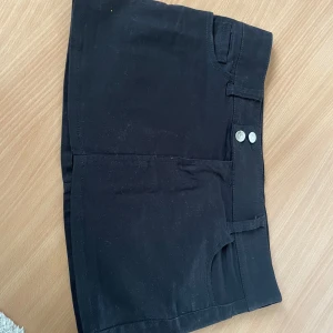 Svarta högmidjade shorts med fickor - Svarta shorts med låg midja och två silverfärgade knappar framtill. Klassisk femficksmodell med två bakfickor och bälteshällor. Snygg och enkel design som passar till många olika stilar. Materialet känns som bomull eller en bomullsblandning.