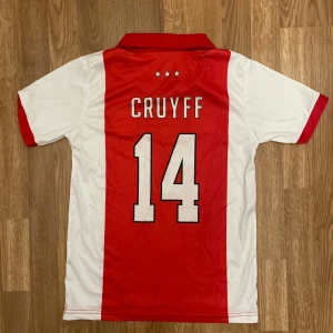Ajax Cruyff 14 retro fotbollströja - Klassisk Ajax fotbollströja med röd mittpanel och vita sidor, Cruyff 14 på ryggen och TDK som sponsor framtill. Ikonisk krage, broderad Ajax-logga och Le Coq Sportif-märke. Perfekt för dig som älskar retrostil och legendariska spelare.
