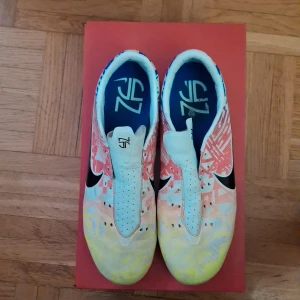 Nike fotbollsskor i färgglad design - Snygga Nike fotbollsskor med unik grafisk design i vitt, gult, blått och rött. Skorna har svart swoosh, perforerad ovandel och FG-dobbar för spel på gräs. Lätta och smidiga med täckt snörning för bättre bollkontroll. Perfekta för dig som vill sticka ut på planen. Snöre medföljer! 