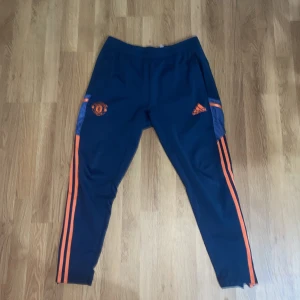 Äkta Manchester United träningsbyxor Adidas - Mörkblå träningsbyxor från Adidas med Manchester United-logga och orangea detaljer längs benen. Byxorna har smal passform, elastisk midja och är tillverkade i polyester. Perfekta för fotbollsträning eller chill.