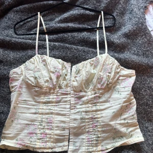 Blommig korsettopp från Gina Tricot - Supersöt korsettopp från Gina Tricot med smala axelband och blommigt mönster i pastelliga toner av rosa och grönt på en ljusbeige bas. Toppen har hysk- och hakknäppning framtill och lätt rynkade kupor. Perfekt för att lyfta vilken outfit som helst!