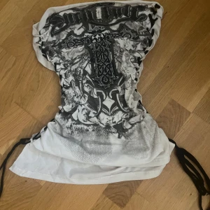 Vit och svart tubtopp med tryck - Cool tubtopp i vitt med svart grafiskt tryck och korsdetalj framtill. Toppen har snörning i sidorna för en justerbar passform. Perfekt för dig som vill sticka ut med edgy stil.