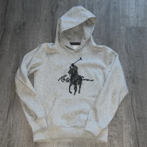 Grå hoodie från Polo Ralph Lauren - Snygg grå hoodie från Polo Ralph Lauren med stor svart polospelare och signatur på bröstet. Klassisk känguruficka, huva och ribbade muddar. Mjuk bomullsblandning som är perfekt för chill dagar. Passar dig som gillar stilren streetwear.