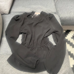 Svart blus med puffärm från ONLY - Svart blus från ONLY med rund halsringning, långa puffärmar och markerad midja med resår. Blusen har lätt volang nertill och stängs med knapp i nacken. Perfekt till både jeans och kjol för en trendig look.