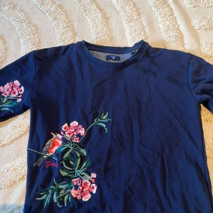 Blå t-shirt med broderi från Gant - Mörkblå t-shirt från Gant med korta ärmar och snyggt broderi av blommor och en fågel på bröstet och ena ärmen. Skön bomullskvalitet och rund halsringning. Passar bra till jeans och kjol