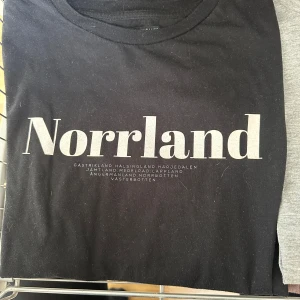 Svart Norrland t-shirt från Sortn Company - Svart t-shirt från Sortn Company med stor vit text 'Norrland' på bröstet och mindre text med norrländska landskap.  Använd fåtal gånger 
