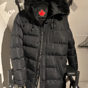 Svart pufferjacka Zavetti Canada - Säljer en svart pufferjacka från Zavetti Canada med stor huva och fuskpäls. Jackan har dragkedja, bröstficka och två sidofickor. Badge på ärmen och snygga detaljer. Perfekt för kalla dagar och riktigt street.