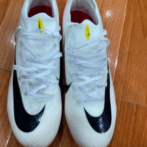 Nike Air Zoom vita fotbollsskor - Snygga vita Nike Air Zoom fotbollsskor med svart swoosh, rosa och röda detaljer på sulan och hälen. Ovandelen är i syntetmaterial med ventilerande mesh och snörning. Perfekta för dig som vill sticka ut på planen med stilrena och sportiga skor.