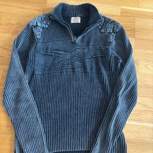 Mörkblå ribbad stickad tröja XL - Ribbstickad grå tröja från Lyjeassion i storlek XL. Tröjan har ett snyggt örnmotiv framtill, halv dragkedja vid halsen och coola axeldetaljer med knappar. Perfekt för dig som gillar unika detaljer och en bekväm passform.