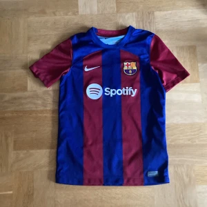 FC Barcelona Nike matchtröja - Säljer en officiell FC Barcelona matchtröja från Nike med Dri-FIT-material. Tröjan har breda blå och vinröda ränder, klubbmärke och sponsortryck på bröstet samt UNHCR-logga på ryggen. Kortärmad och tillverkad i polyester, perfekt för fotbollsälskare. Köpt från Nike’s hemsida så är inte fejk
