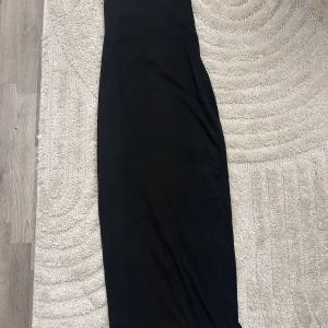 Svart långklänning med snörning - Snygg svart långklänning med tunna axelband och öppen rygg med snörningsdetalj. Klänningen har en slimmad passform och är perfekt för dig som gillar minimalistisk och stilren look. Materialet känns mjukt och följsamt mot kroppen.