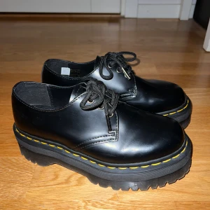 Svarta Dr. Martens med platåsula - Säljer ett par svarta Dr. Martens med kraftig platåsula och klassiska gula sömmar. Skorna har snörning, rund tå och är tillverkade i blankt skinn. Aldrig använda.