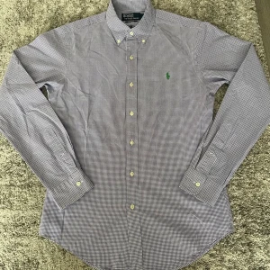 Rutig skjorta från Polo Ralph Lauren - Snygg rutig skjorta från Polo Ralph Lauren i vitt och blått med klassisk button-down krage och grönt broderat logotyp på bröstet. Skjortan har långa ärmar, knappar framtill och är tillverkad i mjuk bomull. Perfekt för en clean och stilren look. Storlek S, hör av dig vid fundering
