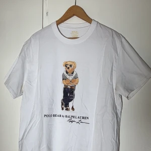 Vit Polo Bear t-shirt från Ralph Lauren - Vit t-shirt från Ralph Lauren med ikoniskt Polo Bear-tryck på bröstet. Modellen är custom slim fit och har klassisk rund hals och korta ärmar. Tillverkad i mjuk bomull, perfekt för en clean och stilren look.