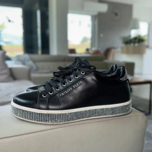 Philipp Plein svarta sneakers med strass - Svarta sneakers i skinn från Philipp Plein med glittrande strassdetaljer runt hela sulan och bak på hälen. Skorna har snörning, metallögla och logga på sidan. Perfekta för dig som vill sticka ut med en lyxig vibe.