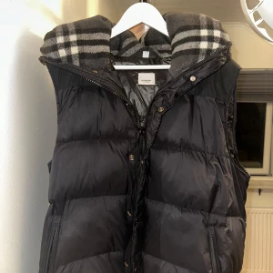 Svart dunväst från Burberry - Snygg svart dunväst från Burberry med klassisk rutig huva i grått och vitt. Västen har dragkedja och knappar framtill samt två fickor med dragkedja. Perfekt för lager-på-lager och ger en cool vibe till din outfit.
