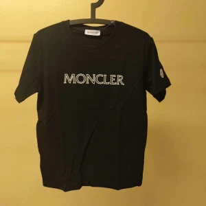 Svart Moncler t-shirt - Svart t-shirt från Moncler med klassisk vit logotyp tryckt på bröstet och broderad patch på ärmen. T-shirten har rund halsringning och korta ärmar. Tillverkad i mjuk bomull för en skön och stilren look.