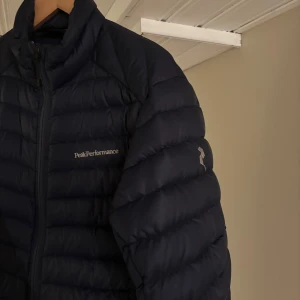 Peak performance jacka - Säljer en mörkblå dunjacka från Peak Performance med diskret logga på bröstet och ärmen. Jackan har quiltad design, dragkedja framtill och hög krage. Perfekt för kyliga dagar och riktigt snygg. Jackan är i jätte fint skick!!!