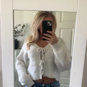 Vit stickad kofta med knyt framtill - Supermysig vit stickad kofta med croppad passform och långa ärmar. Koftan har v-ringning och knyts framtill med två breda snören. Perfekt att slänga över ett linne eller topp för en chill look.