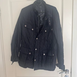 Barbour jacka - Snygg mörkblå quiltad jacka från Barbour International med silverfärgade tryckknappar och klassisk krage. Jackan har flera fickor framtill och Barbour-logga på ärmen. Perfekt för dig som gillar stilren och tidlös design. Har en defekt se bild 4