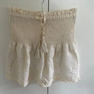 Beige kortkjol med smock och knyt - Supersöt beige kortkjol med smockad midja och knytband med tofsar. Köpte den i Spanien i en Zara butik och är helt oanvänd! Pris kan diskuteras vid snabbköp💕