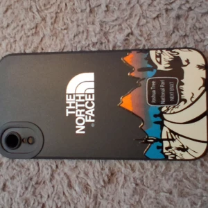 Svart mobilskal The North Face - Snyggt svart mobilskal från The North Face med coolt motiv av berg och natur i orange, blått och beige. Skalet har en matt finish och tryckt logga på baksidan. Passar perfekt för dig som gillar outdoor-vibbar och vill ha ett unikt skal till mobilen.