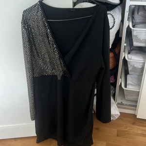 Svart kortklänning med glittrig ärm - Snygg svart kortklänning med lång ärm där ena sidan är täckt av glittriga paljetter i silver. Djup v-ringning framtill och figurnära passform. Perfekt för dig som vill sticka ut med en cool och trendig look.