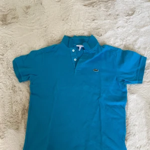 Blå pikétröja från Lacoste - Snygg blå pikétröja från Lacoste med klassisk krage och knappar framtill. Ikoniska krokodilloggan på bröstet.  Pris kan diskuteras💕💕
