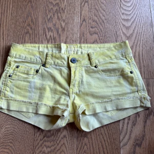 Gula jeansshorts med fransig kant - Snygga gula jeansshorts med fransig kant och slitna detaljer. Klassisk femficksmodell med knapp och dragkedja framtill. Perfekta för varma dagar när du vill sticka ut lite extra. Sköna och coola shorts som ger sommarvibbar.