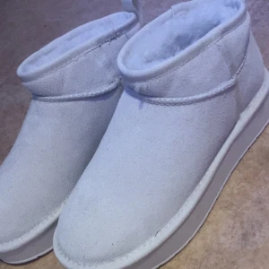 Vita UGG boots i mocka med foder - Säljer ett par vita UGG boots i mocka med fluffigt foder och platt sula. Skorna har en låg, rundad siluett och är perfekta för kalla dagar. Klassisk UGG-design med mjukt material och bekväm passform. Aldrig använda. köptes för 700kr men säljer nu dom för 250.